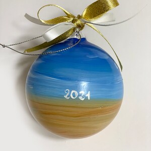 K&ouml;nnte beinhalten: Eine kugelf&ouml;rmige Glasdekoration mit einer handgemalten Landschaft aus blauem Himmel &uuml;ber einem beigefarbenen Sandstrand. Das Jahr 2021 ist in wei&szlig;er Schrift in der Mitte aufgemalt. Ein gold-wei&szlig;es Band ist oben zu einer Schleife gebunden.