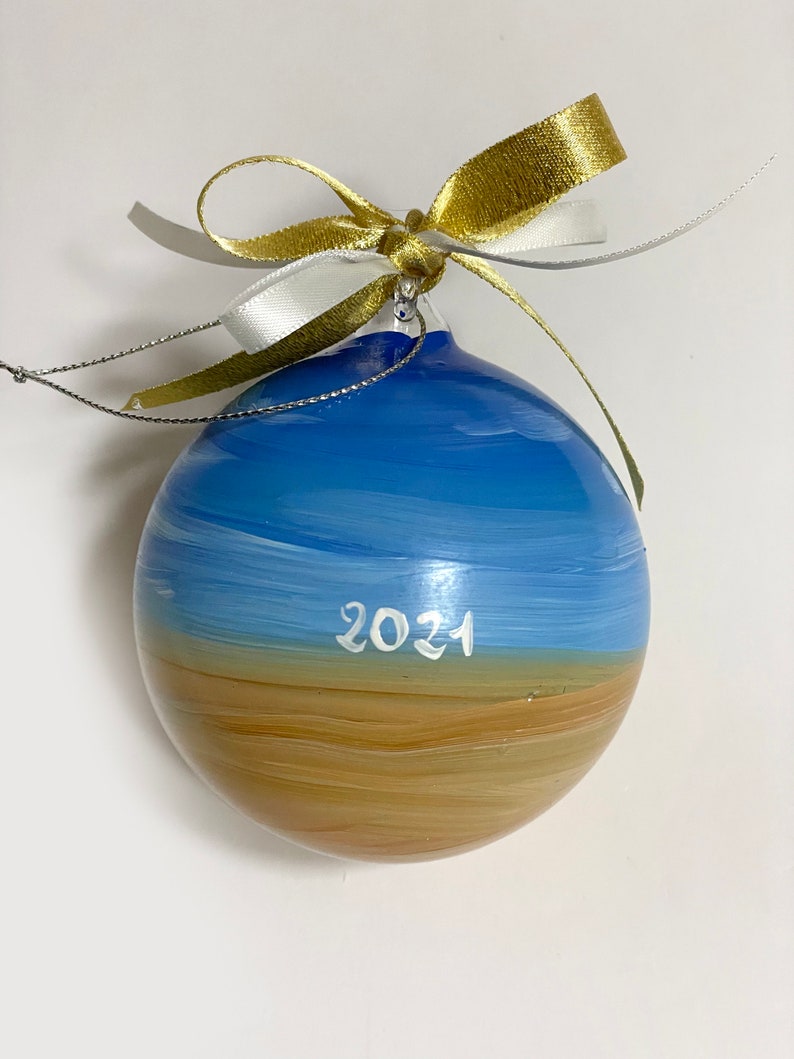 K&ouml;nnte beinhalten: Eine kugelf&ouml;rmige Glasdekoration mit einer handgemalten Landschaft aus blauem Himmel &uuml;ber einem beigefarbenen Sandstrand. Das Jahr 2021 ist in wei&szlig;er Schrift in der Mitte aufgemalt. Ein gold-wei&szlig;es Band ist oben zu einer Schleife gebunden.