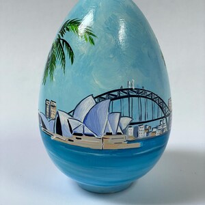 May include: A hand-painted egg featuring a detailed scene of the Sydney Opera House and Harbour Bridge against a light blue sky. The artwork includes green palm fronds in the upper left corner and a deep blue water base, creating a coastal view.