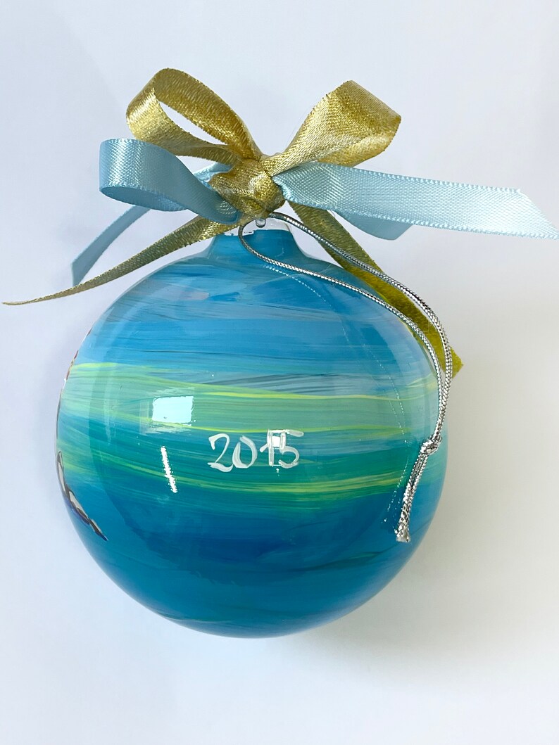 K&ouml;nnte beinhalten: Eine kugelf&ouml;rmige Glasdekoration mit horizontalen Pinselstrichen in Blau, T&uuml;rkis und Gelb. Die Jahreszahl 2015 ist in Wei&szlig; auf der Vorderseite aufgemalt. Ein goldenes und hellblaues Band ist oben befestigt, erg&auml;nzt durch eine silberne Kordel.