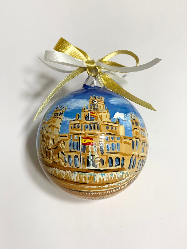 Può includere: Un addobbo natalizio dipinto a mano che raffigura un edificio con una torre dell'orologio, una bandiera spagnola e uno sfondo di cielo blu.