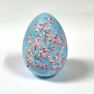 Puede incluir: Un huevo de color azul claro decorado con ramas negras pintadas a mano y racimos de pequeñas flores de cerezo rosas y blancas. La superficie presenta pinceladas visibles en varios tonos de azul, creando una apariencia texturizada en el huevo.
