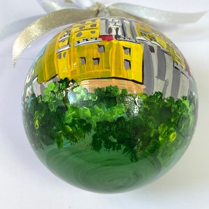 以下が含まれることがあります： 手描きのクリスマスオーナメント。黄色の建物と緑の木々、白いリボンが描かれている。