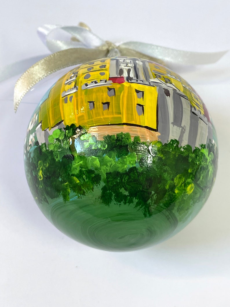 以下が含まれることがあります： 手描きのクリスマスオーナメント。黄色の建物と緑の木々、白いリボンが描かれている。