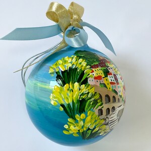 K&ouml;nnte beinhalten: Eine kugelf&ouml;rmige Glasdekoration mit einer handgemalten K&uuml;stenszene, die leuchtend gelbe Blumen im Vordergrund, eine Steinbr&uuml;cke und bunte Geb&auml;ude zeigt. Die Kugel ist mit einem hellblauen Satinband und einer goldenen Metallschleife versehen.