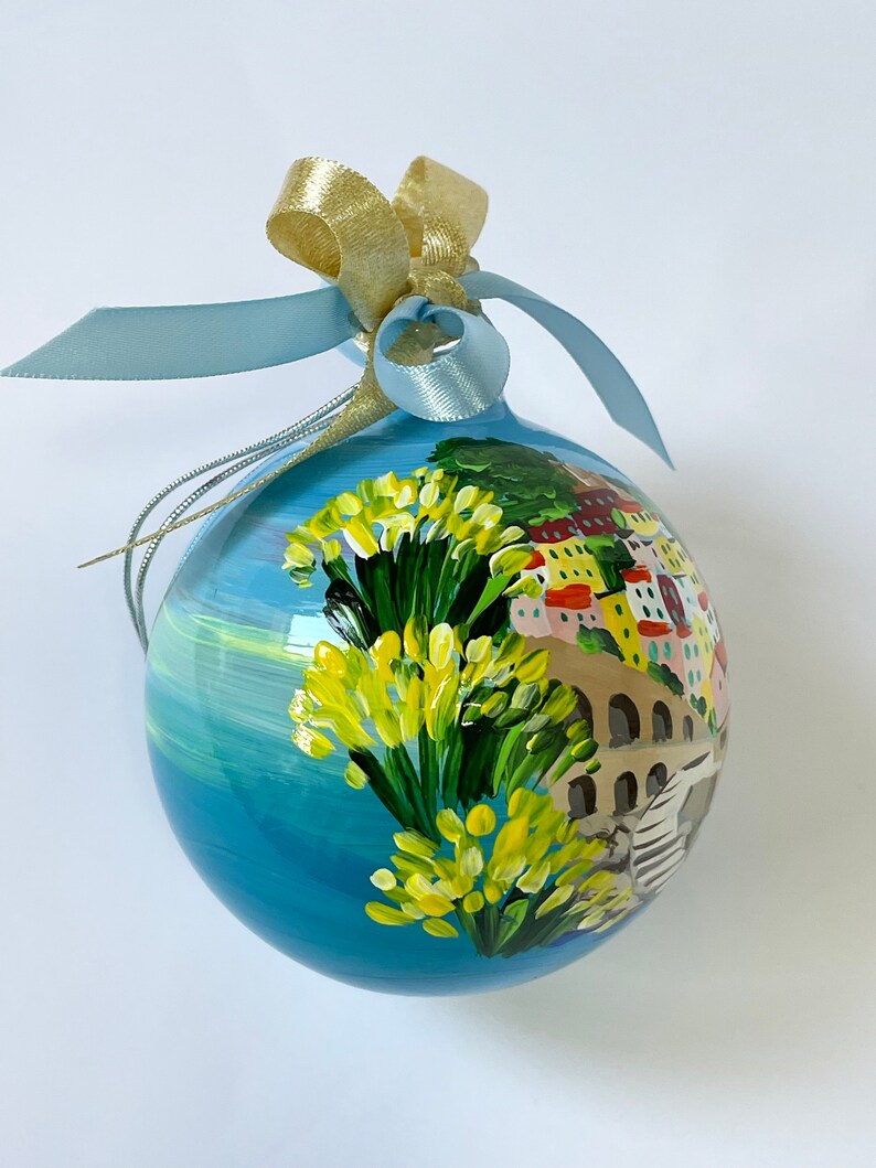 K&ouml;nnte beinhalten: Eine kugelf&ouml;rmige Glasdekoration mit einer handgemalten K&uuml;stenszene, die leuchtend gelbe Blumen im Vordergrund, eine Steinbr&uuml;cke und bunte Geb&auml;ude zeigt. Die Kugel ist mit einem hellblauen Satinband und einer goldenen Metallschleife versehen.