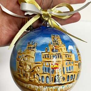 K&ouml;nnte beinhalten: Eine runde Weihnachtskugel aus Glas mit einer handgemalten Szene des Palacio de Cibeles in Madrid, Spanien. Das Geb&auml;ude ist in Beige- und Braunt&ouml;nen vor einem blauen Himmel dargestellt und mit einer gold-wei&szlig;en Schleife zum Aufh&auml;ngen versehen.