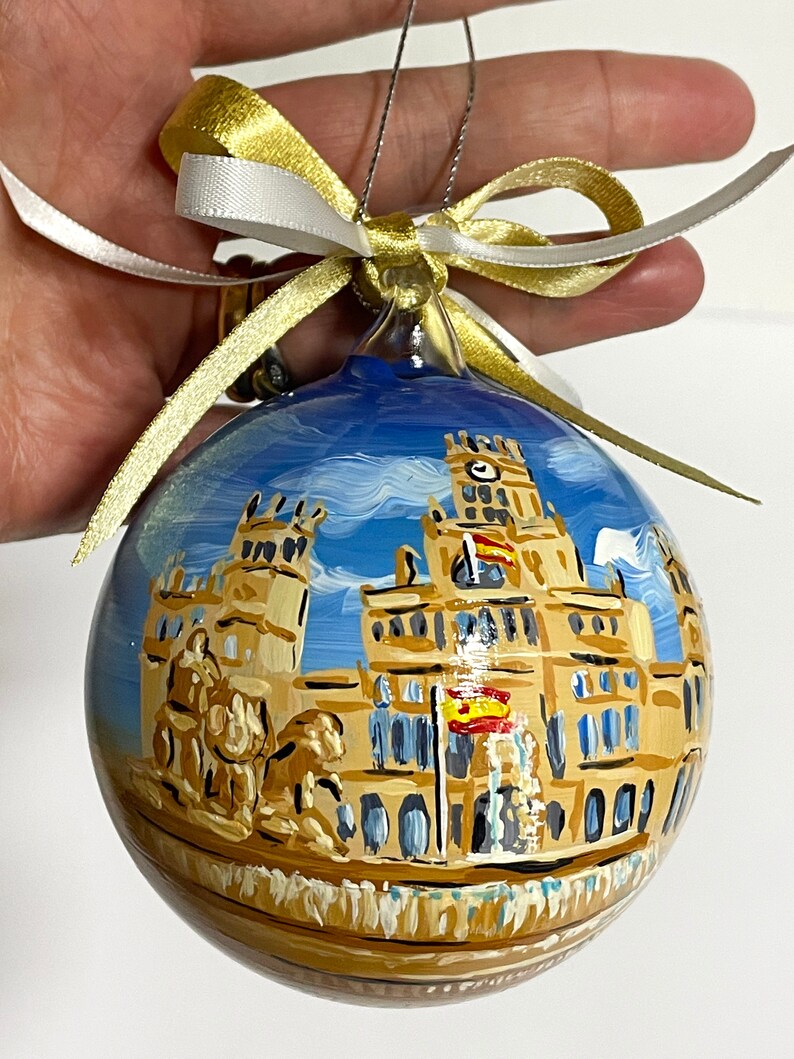 K&ouml;nnte beinhalten: Eine runde Weihnachtskugel aus Glas mit einer handgemalten Szene des Palacio de Cibeles in Madrid, Spanien. Das Geb&auml;ude ist in Beige- und Braunt&ouml;nen vor einem blauen Himmel dargestellt und mit einer gold-wei&szlig;en Schleife zum Aufh&auml;ngen versehen.