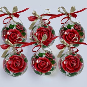 Puede incluir: Un conjunto de seis esferas de cristal transparente, pintadas a mano con rosas rojas vibrantes y hojas verdes. Cada esfera está rematada con un lazo decorativo de cinta dorada y roja brillante, diseñado para colgar como decoración.