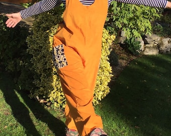 May include: A bright orange jumpsuit with a large patch pocket on the left leg. The pocket has a colorful geometric pattern in shades of blue, yellow, and black. The jumpsuit has a scoop neck and a sleeveless design.