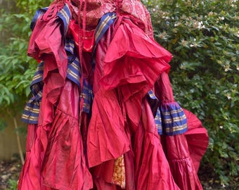 May include: A vibrant red skirt with multiple layers of ruffled fabric. The skirt features accents of blue and gold trim, and a paisley patterned waistband. The skirt is displayed on a mannequin, showcasing its flowing design.