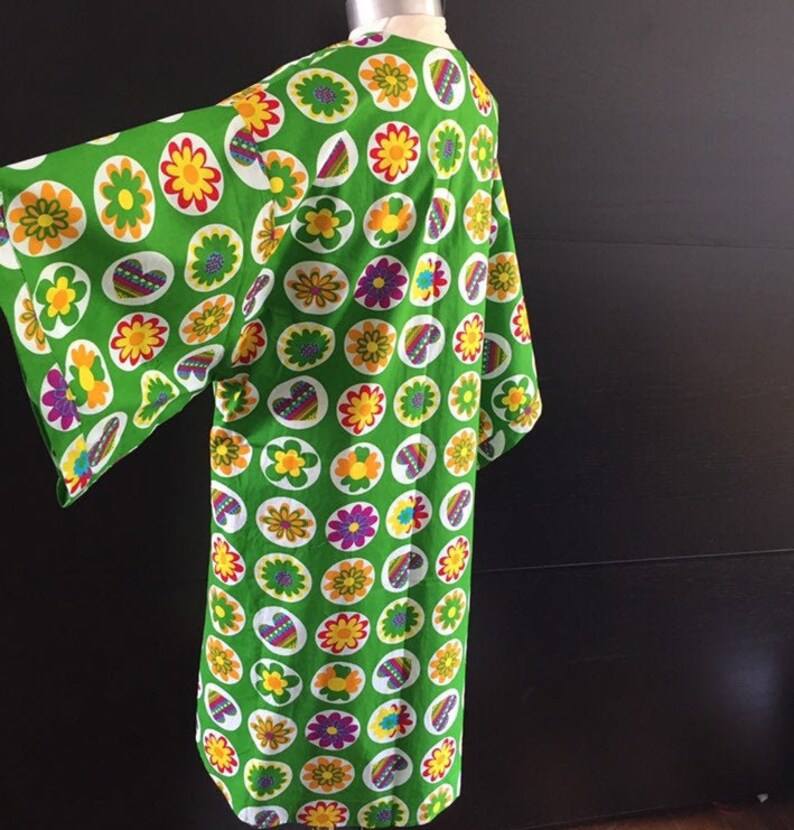 K&ouml;nnte beinhalten: Ein gr&uuml;nes Kimono mit einem bunten Blumenmuster. Das Kimono hat einen weiten, flie&szlig;enden &Auml;rmel und eine lockere Passform.