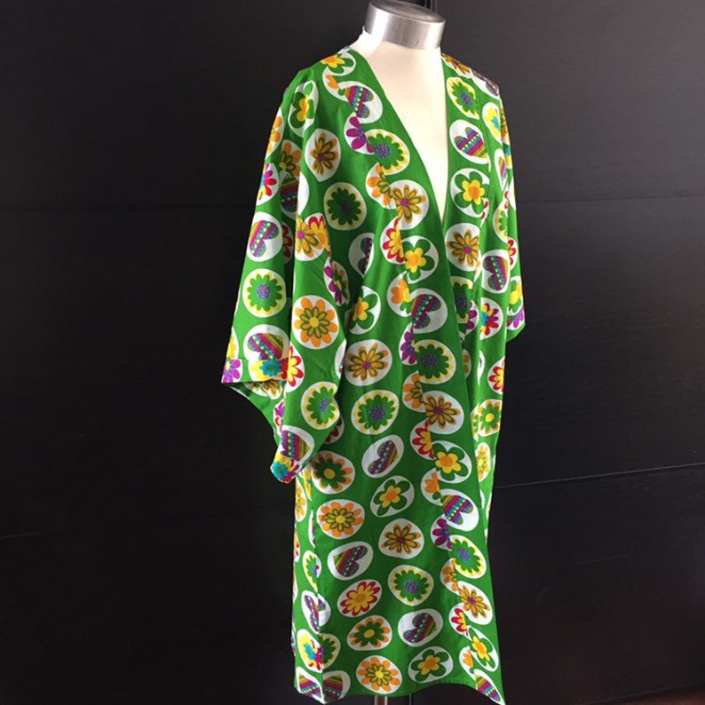 K&ouml;nnte beinhalten: Ein gr&uuml;nes Kimono mit einem farbenfrohen Blumenmuster. Das Kimono hat einen V-Ausschnitt und lange &Auml;rmel. Das Muster zeigt Kreise mit Blumen und Schmetterlingen.