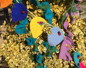 May include: A colourful felt garland featuring leaves and small animals. The garland is made of felt and comes in a variety of colours, including green, blue, yellow, purple and red. The leaves are decorated with small animals, such as birds and ladybugs.