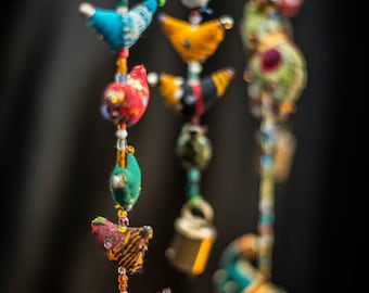 May include: A colorful string of fabric birds with beads and bells hanging from the ceiling. The birds are made of different fabrics and patterns, and they are all strung together with a thin cord. The birds are a variety of colors, including red, blue, green, yellow, and orange.