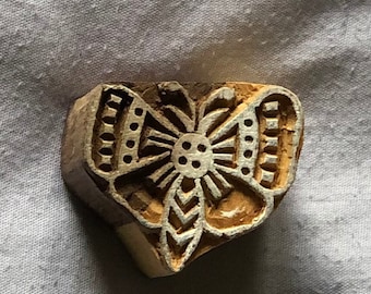 May include: A wooden block printing stamp with a carved butterfly design. The butterfly has a detailed pattern with a central circle and a leaf-like shape at the bottom.