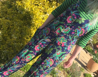 May include: A blue jumpsuit with a floral pattern in shades of pink, green, and gold. The jumpsuit has a wide leg and a fitted bodice. The person is wearing a green and white striped shirt underneath.