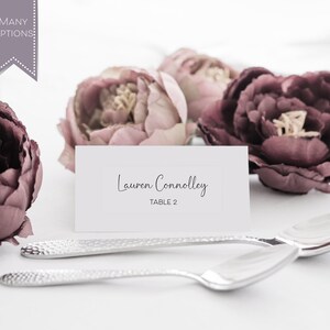 May include: White place card with the name "Lauren Connolly" and "Table 2" printed in black text. The place card is surrounded by pink and burgundy artificial flowers.
