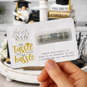 May include: A graduation gift card with a rolled-up wad of cash in a clear plastic holder. The card has gold text that reads "The tassel was worth the hassle" with firework and graduation cap graphics. The card also has text that reads "Spend this wisely or blow it on DoorDash at 2am...either way you earned it!"