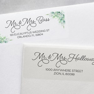 May include: Two white return address labels with black calligraphy text and a green floral design. The top label reads "Mr. & Mrs. Bass" and "123 Eucalyptus Wedding St Orlando, FL 32804". The bottom label reads "Mr. & Mrs. Halloway" and "1000 Anywhere Street Zion, IL 60099".