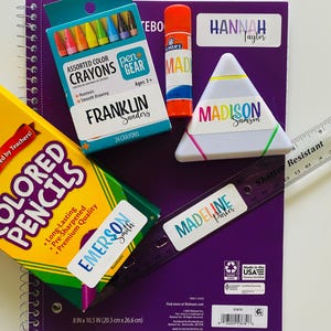 Pu&ograve; includere: Una collezione di materiale scolastico che include un quaderno a spirale viola, una scatola di matite colorate Crayola, una scatola di pastelli a cera Pen+Gear, una colla stick Elmer's, un astuccio triangolare bianco e un righello di 30 cm.