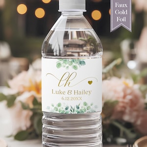May include: A clear plastic water bottle with a white label featuring green eucalyptus leaf illustrations at the top and bottom. The label displays the gold script initials LH, the names Luke & Hailey, and the date 6.12.20XX in gold text.