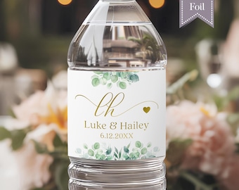 May include: A clear plastic water bottle with a white label featuring green eucalyptus leaf illustrations at the top and bottom. The label displays the gold script initials LH, the names Luke & Hailey, and the date 6.12.20XX in gold text.