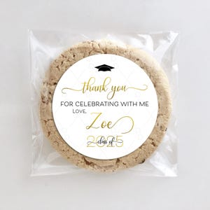 May include: White round sticker with black and gold text. The sticker says "Thank you for celebrating with me. Love, Zoe Class of 2025". A black graduation cap icon is above the text. The sticker is on a brown cookie.