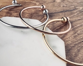 May include: Three simple bangle bracelets with ball ends. The bracelets are silver, rose gold, and gold.