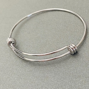 May include: A silver-colored expandable bangle bracelet. The bracelet features a simple design with two decorative knots on either side. The bracelet is made of a shiny, metallic material and is shown against a gray background.