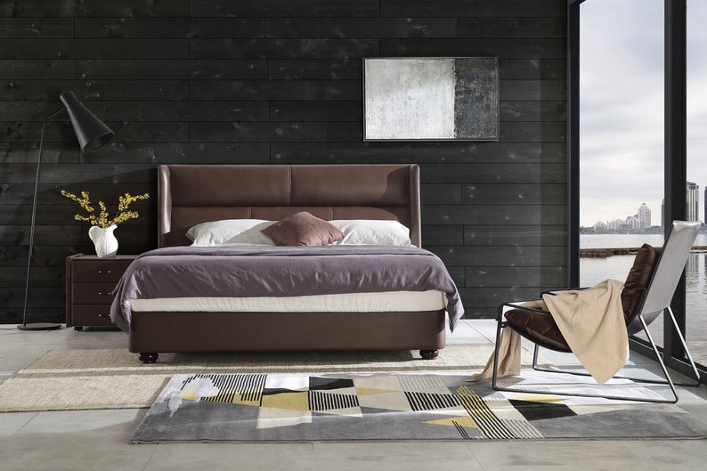 Puede incluir: Una cama con estructura de cuero marr&oacute;n con cabecero y piecero. La cama est&aacute; hecha con un edred&oacute;n morado y almohadas blancas. Una l&aacute;mpara de pie negra est&aacute; en la esquina de la habitaci&oacute;n. Un cuadro abstracto en blanco y negro cuelga de la pared detr&aacute;s de la cama. Una silla marr&oacute;n con una manta beige est&aacute; en primer plano. La habitaci&oacute;n tiene un suelo de madera oscura y una alfombra con un patr&oacute;n geom&eacute;trico.