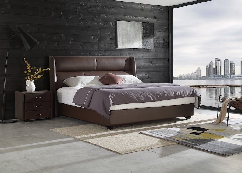 Puede incluir: Una cama con estructura de cuero marr&oacute;n con cabecero y piecero. La cama est&aacute; hecha con ropa de cama blanca y morada y una almohada marr&oacute;n. La cama est&aacute; en un dormitorio con una pared de madera oscura y una ventana con vista a un horizonte urbano.