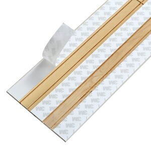 Puede incluir: Primer plano de una tira adhesiva blanca con moldura de madera. La tira adhesiva tiene el texto "3M" impreso en ella.