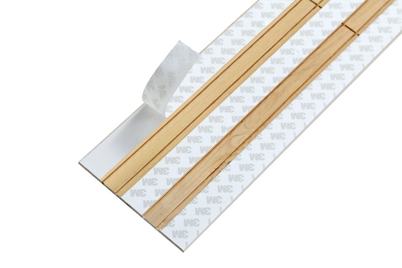 Puede incluir: Primer plano de una tira adhesiva blanca con moldura de madera. La tira adhesiva tiene el texto "3M" impreso en ella.