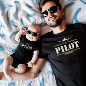 May include: A father and son wearing matching black t-shirts with the words "Pilot" and "Co-Pilot" printed on them. The father is wearing sunglasses and the son is wearing sunglasses. The shirts feature a small airplane graphic.