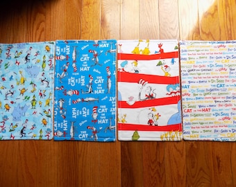 Könnte beinhalten: Vier Stoff-Tischsets mit Illustrationen aus Büchern von Dr. Seuss. Die Tischsets zeigen Charaktere und Text aus "Der Kater mit Hut" und anderen Seuss-Geschichten. Farben sind Blau, Rot, Weiß und Gelb.
