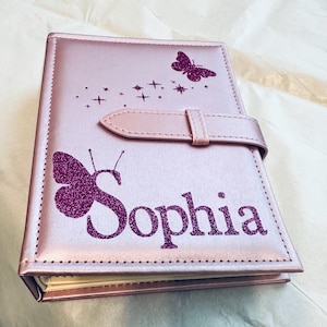 May include: A pink leather-bound journal with a butterfly design and the name "Sophia" in glitter lettering. The journal has a strap closure and a glittery butterfly design.