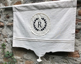 May include: A white fabric banner with a black wreath and the letter "R" in the center. The banner is hanging from a rod and has a white tassel at the bottom.