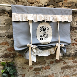 May include: A gray fabric valance with cream-colored ruffles and ties. The valance has a vintage-style sign that reads "FRESH SOAP AND WATER HAND TOWELS FREE 5¢". The valance is hanging on a white rod against a brick wall.