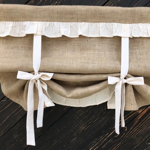 Puede incluir: Una cortina romana de arpillera marrón con ribete blanco con volantes y lazos de tela blanca. La cortina está colgada de una varilla de madera y está sobre un fondo de madera oscura.