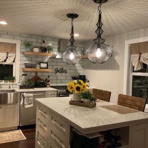 Pu&ograve; includere: Un'isola da cucina bianca con un piano di lavoro bianco e un lavello in stile farmhouse. L'isola ha un piano in legno e una base bianca con cassetti. Ci sono due luci a sospensione nere appese sopra l'isola. La cucina ha un backsplash in piastrelle bianche del metro e un cartello che dice "Gather".