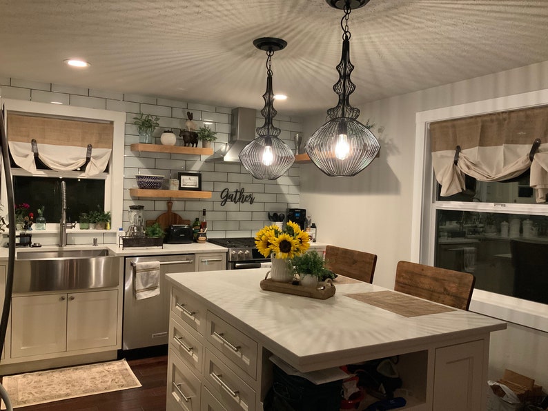 Pu&ograve; includere: Un'isola da cucina bianca con un piano di lavoro bianco e un lavello in stile farmhouse. L'isola ha un piano in legno e una base bianca con cassetti. Ci sono due luci a sospensione nere appese sopra l'isola. La cucina ha un backsplash in piastrelle bianche del metro e un cartello che dice "Gather".