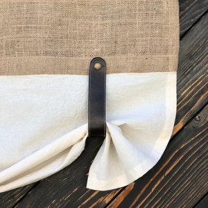 Pu&ograve; includere: Un fermacortina in pelle marrone con un bottone in ottone, che tiene indietro una tenda di lino bianco su uno sfondo di legno scuro.