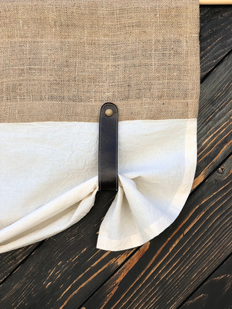 Pu&ograve; includere: Un fermacortina in pelle marrone con un bottone in ottone, che tiene indietro una tenda di lino bianco su uno sfondo di legno scuro.