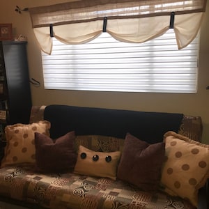 Pu&ograve; includere: Un divano futon marrone con una trapunta trapuntata e diversi cuscini. I cuscini sono marroni, beige e beige chiaro con pois e bottoni decorativi. Il futon &egrave; davanti a una finestra con una bordura bianca e un top in juta marrone.