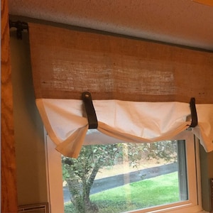 Pu&ograve; includere: Una tenda a valance bianca in tessuto con lacci in pelle marrone &egrave; appesa sopra una tenda a valance in juta. La tenda &egrave; appesa a un'asta di metallo. La finestra dietro la tenda mostra una vista di un albero e erba.