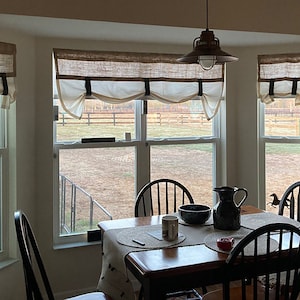 Pu&ograve; includere: Una sala da pranzo con una finestra a bow-window con tre finestre con tende in juta e bianche. Le finestre si affacciano su un campo erboso con una recinzione in legno. Un tavolo in legno con un runner in juta si trova davanti alla finestra. Ci sono quattro sedie nere intorno al tavolo. Un'anfora e una ciotola neri sono sul tavolo.