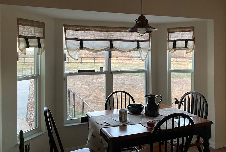 Pu&ograve; includere: Una sala da pranzo con una finestra a bow-window con tre finestre con tende in juta e bianche. Le finestre si affacciano su un campo erboso con una recinzione in legno. Un tavolo in legno con un runner in juta si trova davanti alla finestra. Ci sono quattro sedie nere intorno al tavolo. Un'anfora e una ciotola neri sono sul tavolo.