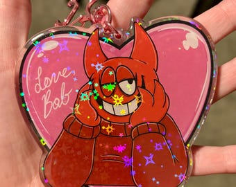 May include: A red heart-shaped keychain with a cartoon character with horns and a red sweater. The character is smiling and has a pink background with the text "love bab" on the left side. The keychain has a holographic finish with glitter.