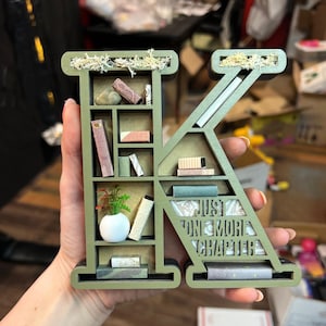 K&ouml;nnte beinhalten: Ein gr&uuml;ner Holzbuchstabe K in Form eines B&uuml;cherregals mit Miniaturb&uuml;chern, einer Pflanze und einer wei&szlig;en Vase. Der Buchstabe tr&auml;gt den Text "Just One More Chapter" an der Seite.